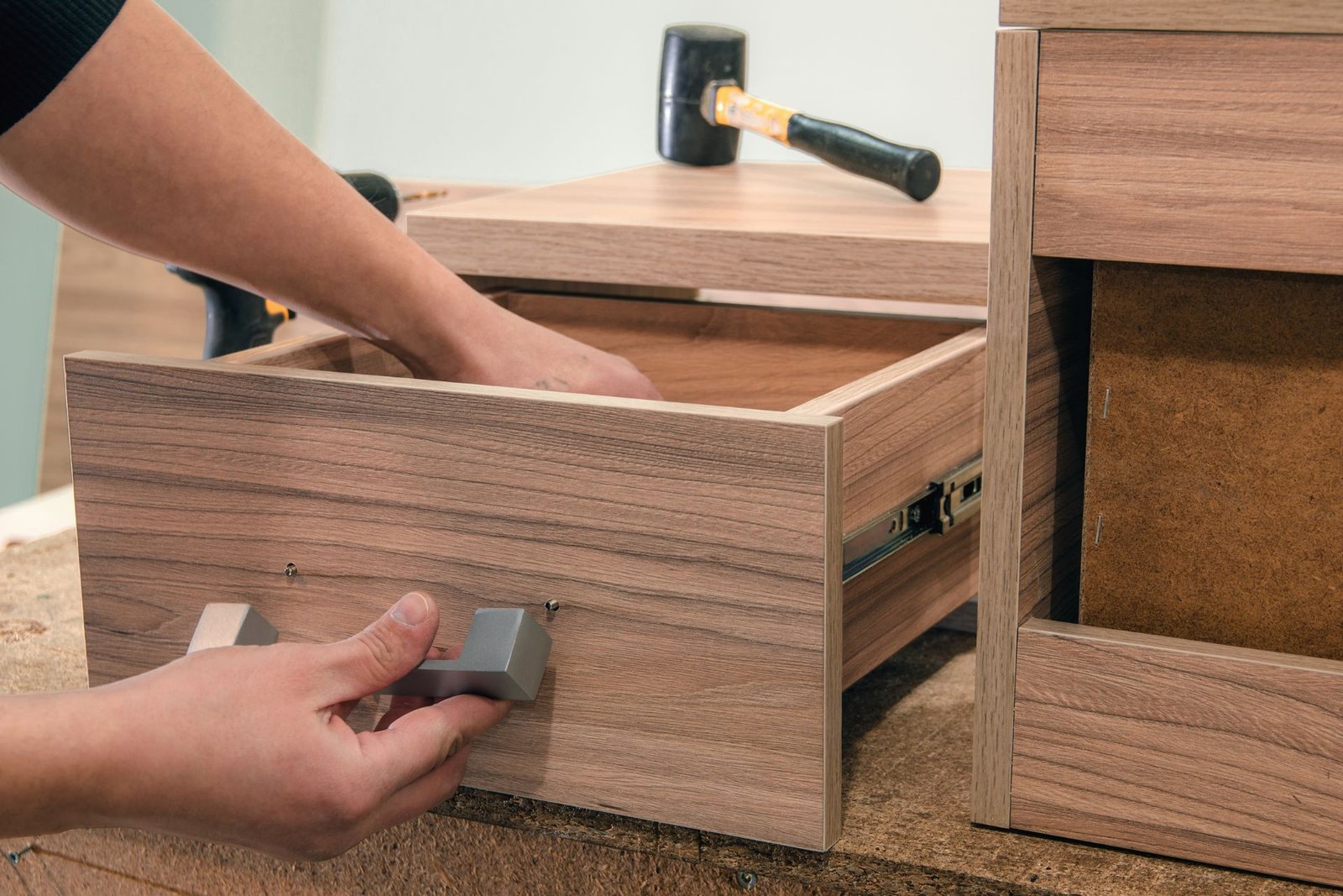 A person installs a handle on a wooden drawer inside a workshop, with various tools visible in the background