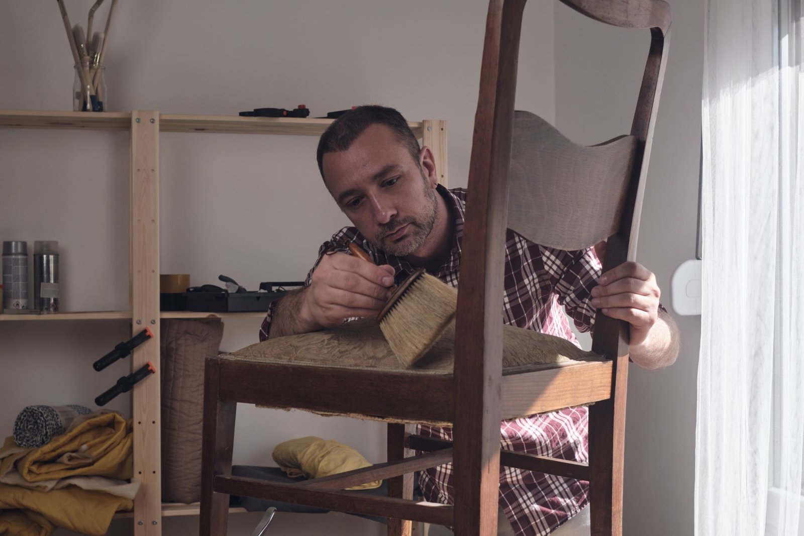 A person repairs a wooden chair in a well-lit room, surrounded by tools and materials for upholstery.
