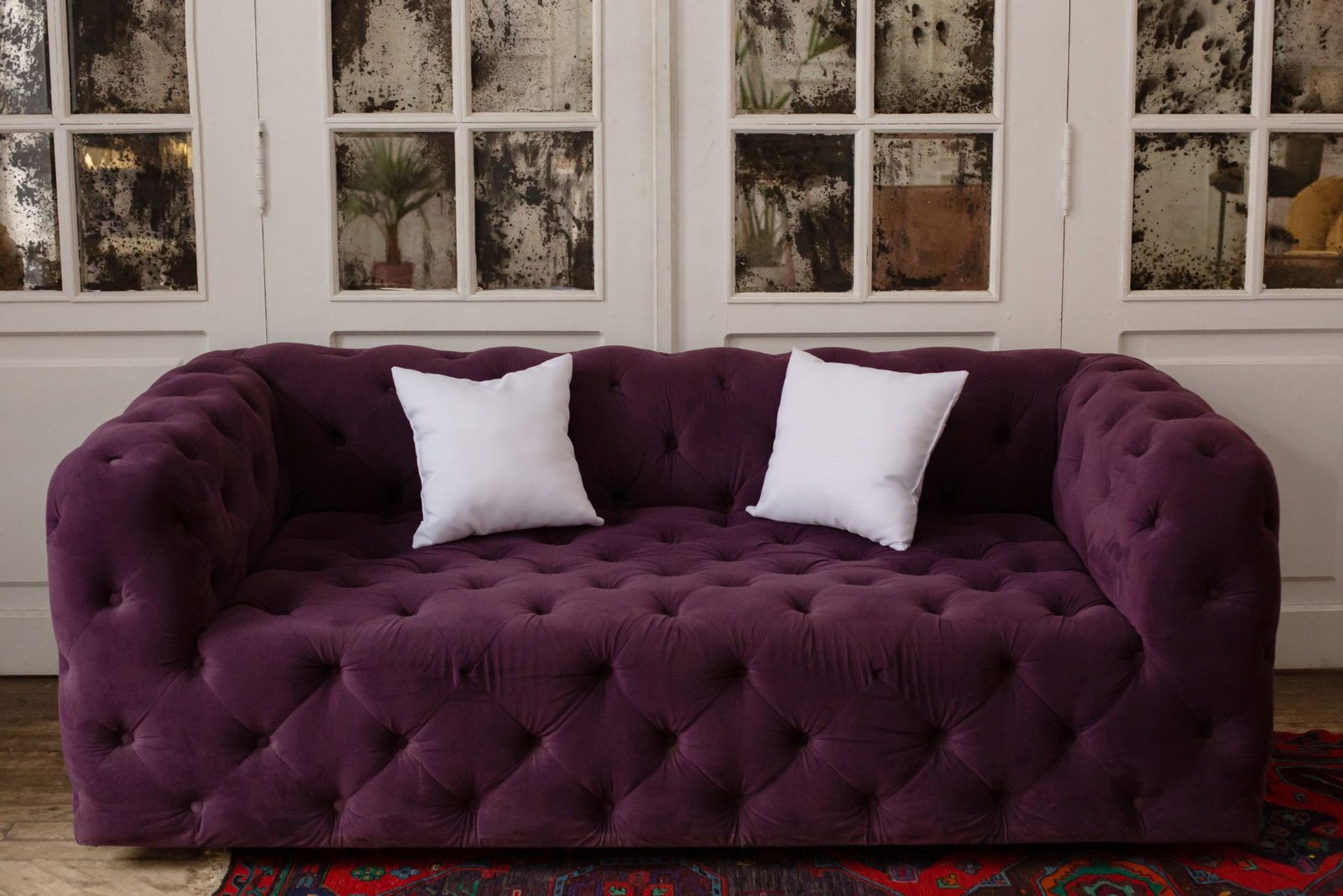 A plush, tufted purple sofa with two white decorative pillows, positioned in front of a window with vintage glass panes.