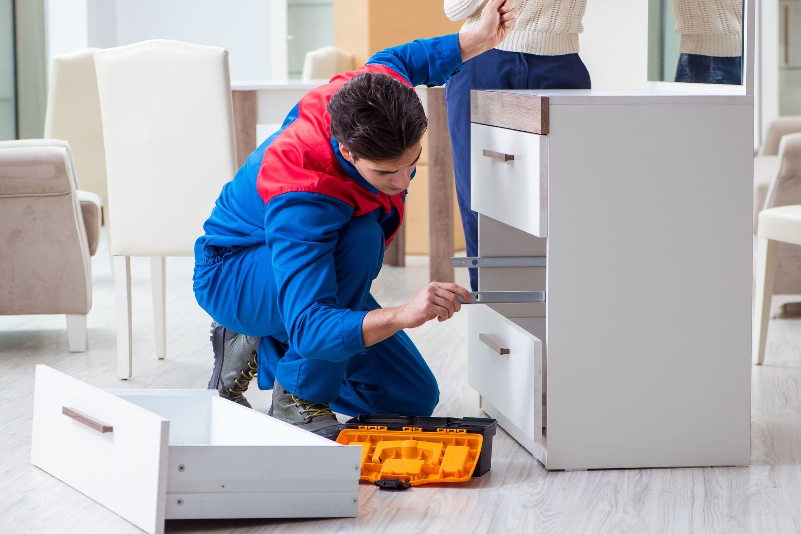 A worker in blue overalls repairs a white dresser, using tools from an open toolbox while adjusting a drawer in a bright room.