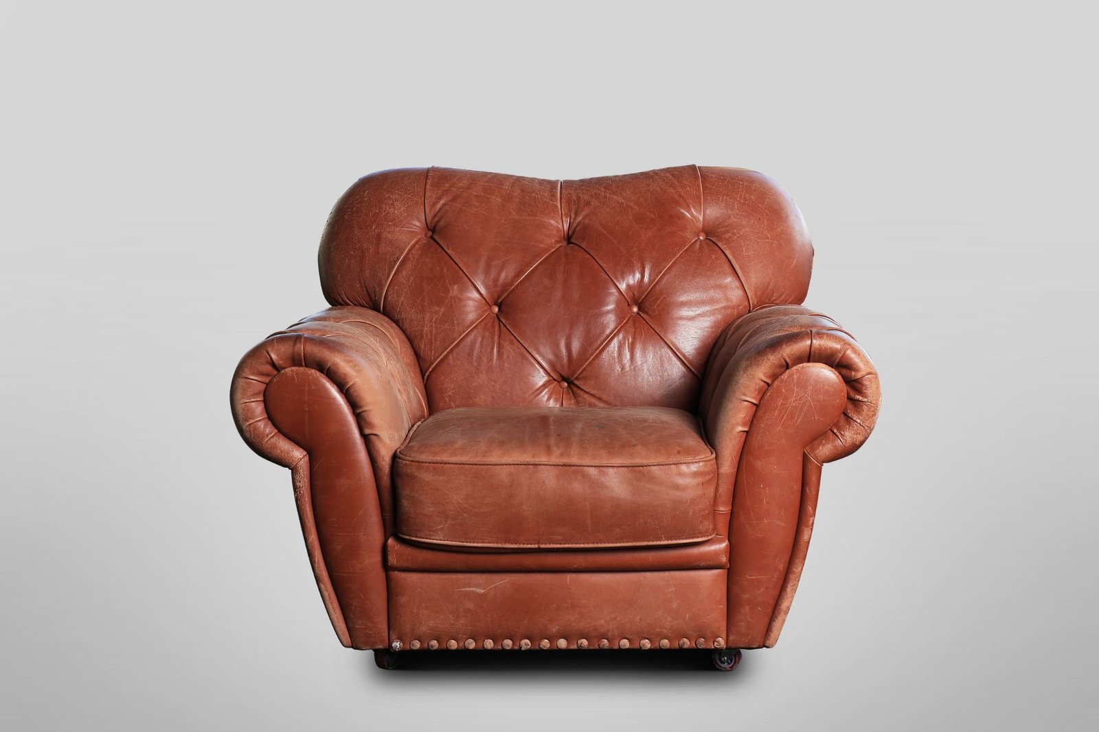 A vintage brown leather armchair with tufted back and rounded arms against a gray background.